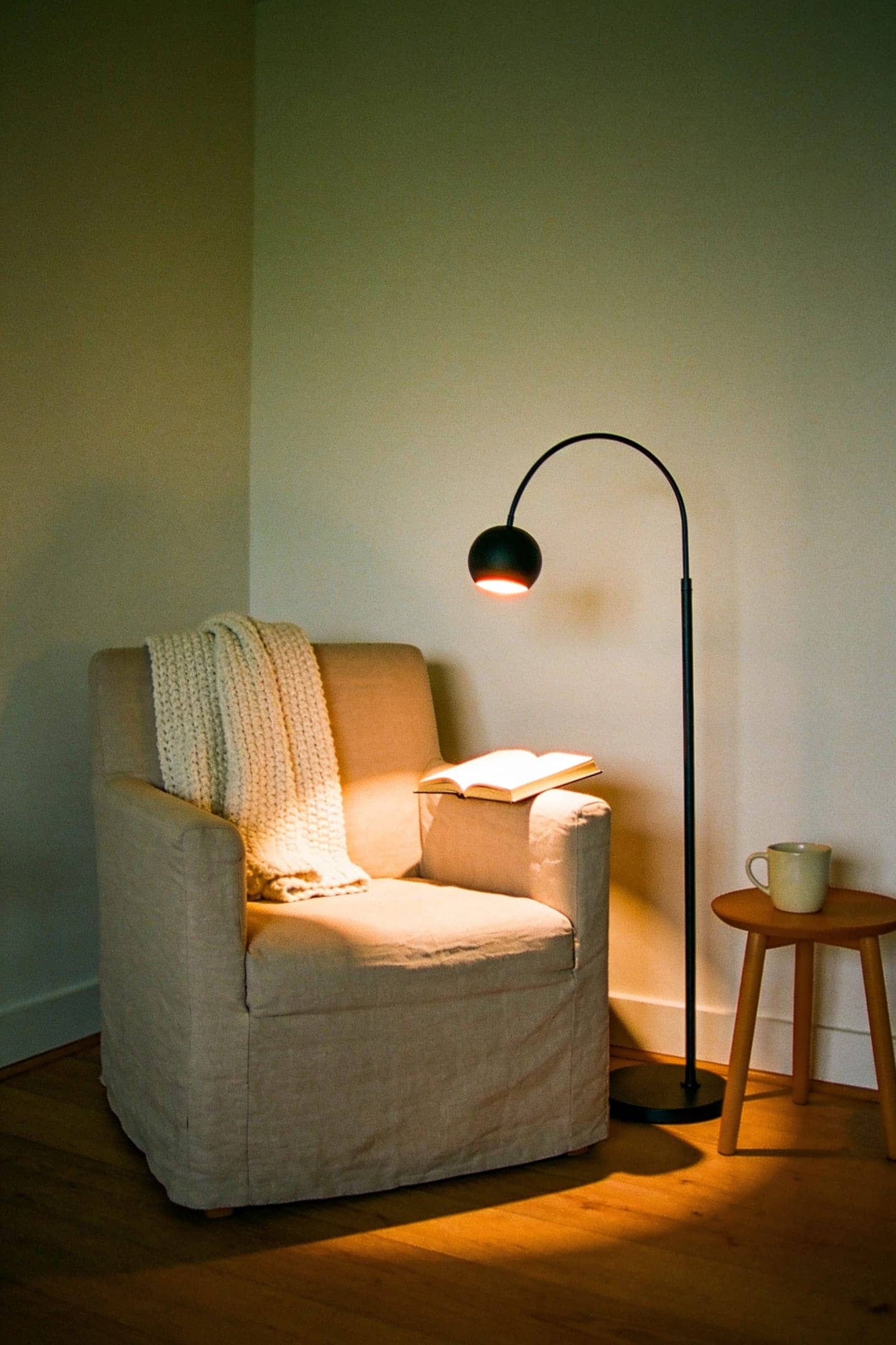 black gooseneck floor lamp bent over a linen armchair, its warm light falling on an open book